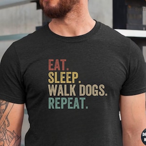 May include: A dark grey heather t-shirt with the text "Eat. Sleep. Walk Dogs. Repeat." in a vintage style font. The text is in a rainbow color scheme.