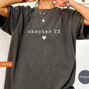 May include: A charcoal grey short-sleeve t-shirt featuring the white text "chapter 23" centred on the chest above a small white heart icon. The garment has a relaxed fit with a crew neckline and a washed, vintage-style fabric texture.