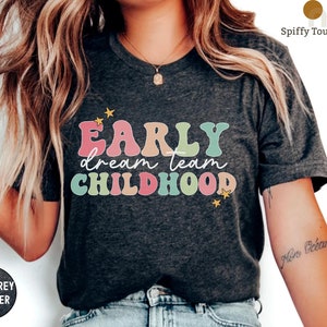 Puede incluir: Una camiseta de color gris oscuro jaspeado con el texto "Early dream team childhood" en letras de colores de estilo retro. Dos peque&ntilde;as estrellas amarillas est&aacute;n a cada lado del texto.