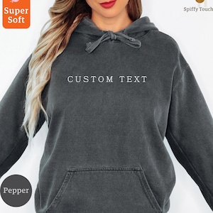Puede incluir: Una sudadera con capucha gris oscuro con el texto "CUSTOM TEXT" en el pecho. La sudadera tiene una capucha con cordón y un bolsillo delantero. La imagen también incluye las palabras "Super Soft" y "Pepper".