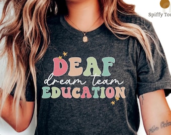 May include: A dark grey heather t-shirt with the text "DEAF dream team EDUCATION" in colorful, retro-style lettering. The text is surrounded by small yellow stars.