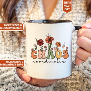 Könnte beinhalten: Eine weiße Keramiktasse mit schwarzem Rand und Henkel. Die Tasse hat ein Blumenmuster mit dem Text "CHAOS coordinator" in bunten Buchstaben.