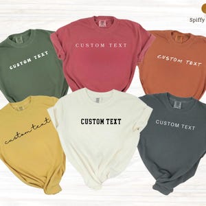 May include: Six colorful t-shirts in various shades, including green, red, and yellow. Each shirt features the words "CUSTOM TEXT" printed across the front. The shirts are displayed on a light wood-grain surface.