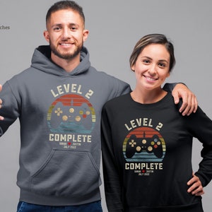 Puede incluir: Dos sudaderas con capucha grises y negras con un gráfico de mando de videojuego retro y el texto "Nivel 2 Completo" con una fecha y nombres.