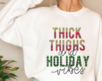 Puede incluir: Una sudadera blanca con el texto "THICK THIGHS and HOLIDAY vibes". El texto combina estampado de leopardo, cuadros rojos y cuadros verdes. Una prenda casual y festiva.