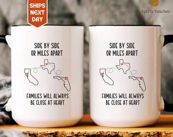 May include: Two white ceramic mugs with black handles. The mugs feature a design of three states, California, Texas, and Florida, with hearts and dotted lines connecting them. The text on the mugs reads "Side by side or miles apart, families will always be close at heart."