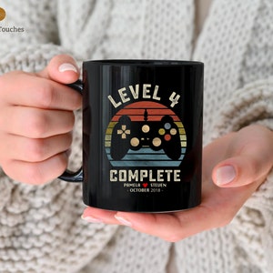 May include: Black ceramic mug with a retro-style design featuring a video game controller and the text "Level 4 Complete". The mug also includes the names "Pamela" and "Steven" and the date "October 2018".