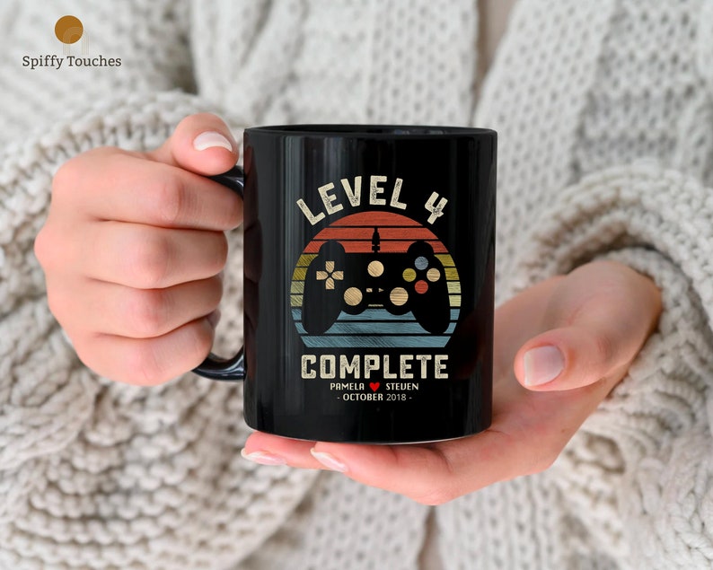 May include: Black ceramic mug with a retro-style design featuring a video game controller and the text "Level 4 Complete". The mug also includes the names "Pamela" and "Steven" and the date "October 2018".
