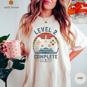 Op de afbeelding: Een wit T-shirt met een retro-stijl grafische afbeelding van een videogamecontroller en de tekst "Level 2 voltooid" met de namen "Kyle" en "Molly" en de datum "November 2020".