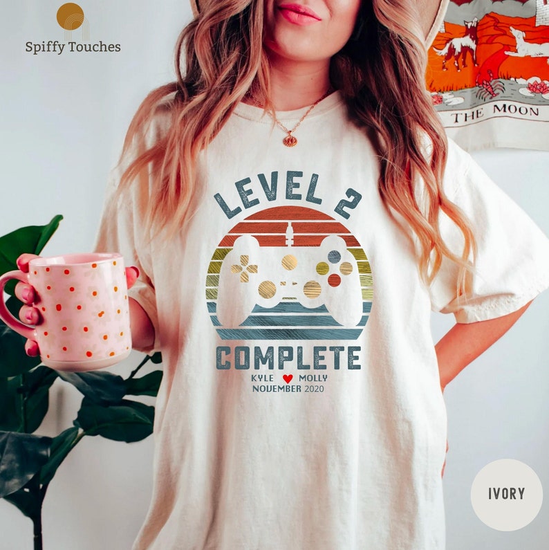 Op de afbeelding: Een wit T-shirt met een retro-stijl grafische afbeelding van een videogamecontroller en de tekst "Level 2 voltooid" met de namen "Kyle" en "Molly" en de datum "November 2020".