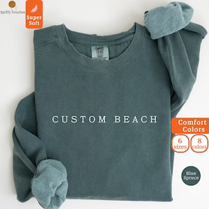 以下が含まれることがあります： 胸元に白い文字でCUSTOM BEACHとプリントされた、ブルー・スプルース色のクルーネック・スウェットシャツ。リブ編みの襟、リブ編みの袖口が付いた長袖、首の内側にComfort Colorsのブランドラベルが施されています。