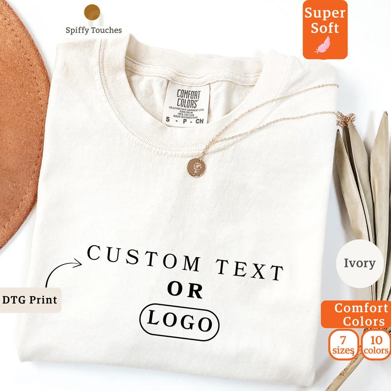 May include: An ivory-coloured t-shirt with the text "CUSTOM TEXT OR LOGO" printed in black. The shirt has a crew neck and short sleeves. A gold necklace with a pendant rests on the shirt. The shirt is labelled as "Super Soft" and "Comfort Colors".