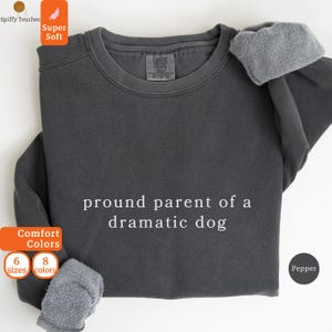 May include: Pepper-colored sweatshirt with the text "pround parent of a dramatic dog" in white. The sweatshirt has a crew neck and long sleeves with a soft, gray lining at the cuffs. The image includes the Comfort Colors and Spiffy Touches logos.