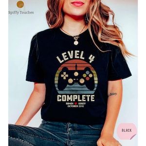 Op de afbeelding: Zwart T-shirt met een retro-stijl videogamecontroller-afbeelding. De tekst "Level 4 Complete" is op het shirt gedrukt, samen met de namen "Randi & Kasey" en de datum "Oktober 2018".