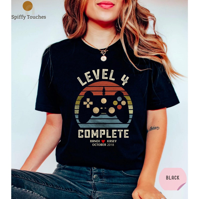 Op de afbeelding: Zwart T-shirt met een retro-stijl videogamecontroller-afbeelding. De tekst "Level 4 Complete" is op het shirt gedrukt, samen met de namen "Randi & Kasey" en de datum "Oktober 2018".
