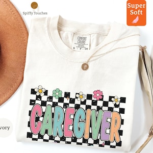 May include: Ivory-colored t-shirt with the word "CAREGIVER" in colorful, retro-style lettering. The design includes a black and white checkered background and daisy accents. The shirt is labeled "Super Soft."