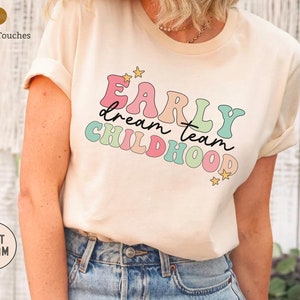 Puede incluir: Una camiseta de color crema con el texto "Early dream team childhood" en colores arco&iacute;ris con estrellas.