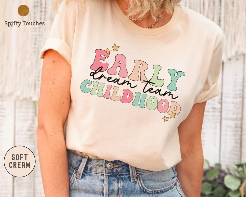 Puede incluir: Una camiseta de color crema con el texto "Early dream team childhood" en colores arco&iacute;ris con estrellas.