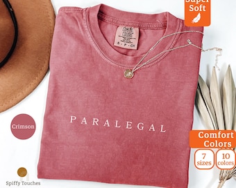 May include: Crimson-colored t-shirt with the word "PARALEGAL" in white lettering. The shirt has a crew neck and is adorned with a delicate gold necklace. The image also shows a brown hat and text that says "Comfort Colors".