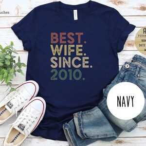 Puede incluir: Una camiseta azul marino con el texto "BEST. WIFE. SINCE. 2010." en una fuente de estilo vintage. El texto est&aacute; en tonos de rojo, naranja, amarillo y verde.