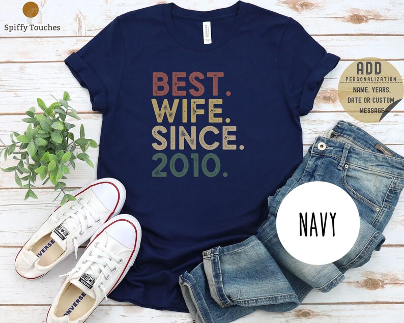 Puede incluir: Una camiseta azul marino con el texto "BEST. WIFE. SINCE. 2010." en una fuente de estilo vintage. El texto est&aacute; en tonos de rojo, naranja, amarillo y verde.