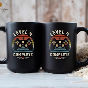 May include: Two black coffee mugs with a retro-style video game controller graphic and the text "Level 4 Complete" with a date and names.