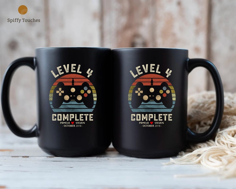 May include: Two black coffee mugs with a retro-style video game controller graphic and the text "Level 4 Complete" with a date and names.