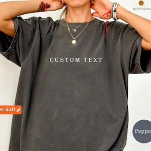 Puede incluir: Camiseta gris oscuro con el texto "CUSTOM TEXT" en blanco. La camiseta tiene mangas cortas y un corte holgado. La imagen también incluye joyas y un tatuaje en el brazo.