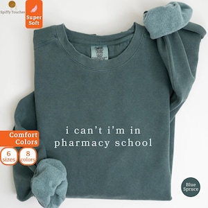 May include: A blue spruce sweatshirt with the text "i can't i'm in pharmacy school" in white. The sweatshirt has a crew neck and long sleeves. The sleeves are cuffed. The image also includes the words "Comfort Colors", "Super Soft", and "6 sizes 8 colors".