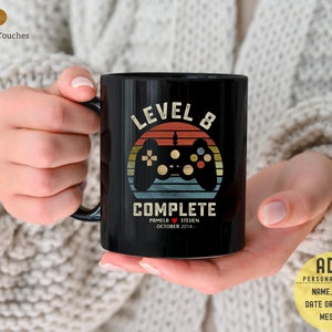 Op de afbeelding: Zwarte koffiemok met een retro-ontwerp met een videogamecontroller en de tekst "Level 8 Complete". De mok bevat ook de namen "Pamela" en "Steven" en de datum "Oktober 2014".