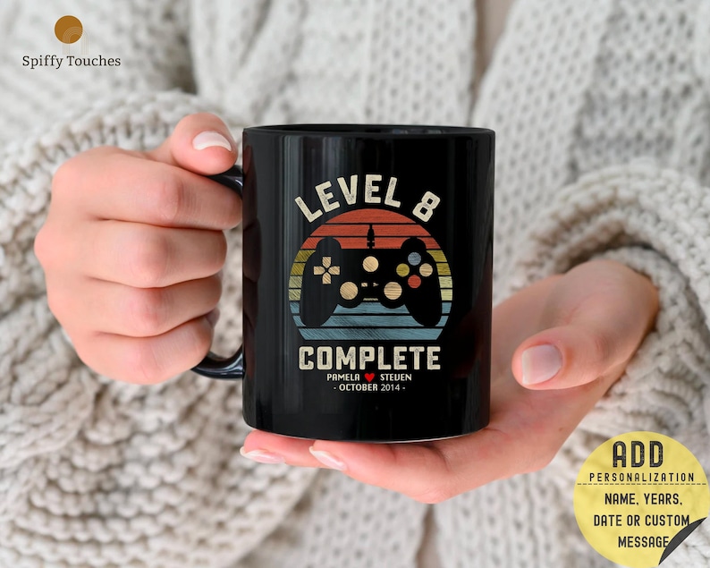 Op de afbeelding: Zwarte koffiemok met een retro-ontwerp met een videogamecontroller en de tekst "Level 8 Complete". De mok bevat ook de namen "Pamela" en "Steven" en de datum "Oktober 2014".