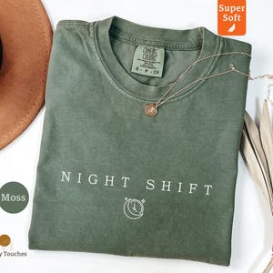 Puede incluir: Una camiseta verde musgo con las palabras "NIGHT SHIFT" impresas en blanco. Un collar dorado con un colgante circular descansa sobre la camiseta. La camiseta está doblada y colocada sobre una superficie blanca.