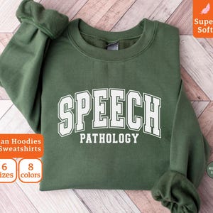 Puede incluir: Sudadera verde militar con las palabras "SPEECH PATHOLOGY" en letras blancas. La sudadera es una sudadera con capucha Gildan y está disponible en 6 tallas y 8 colores. La imagen también incluye las palabras "Super Soft".