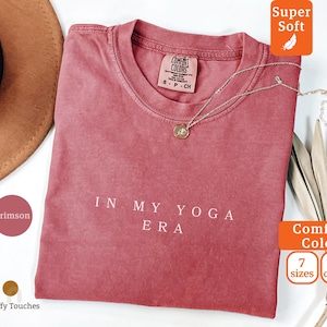 May include: Crimson-colored t-shirt with the text "IN MY YOGA ERA" in white. The shirt has a crew neck and is made of a soft material. A gold necklace with a pendant is draped across the shirt. The shirt is from Comfort Colors.