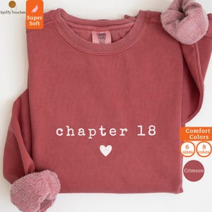 Puede incluir: Sudadera de color carmesí con el texto "chapter 18" y un corazón blanco. La sudadera tiene cuello redondo y mangas largas con un forro suave y difuso en los puños. Se ve la marca "Comfort Colors".
