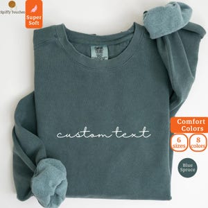 Puede incluir: Una sudadera azul abeto con cuello redondo con las palabras "custom text" en escritura blanca. La sudadera tiene una etiqueta que dice "Comfort Colors". La imagen también incluye texto que dice "Super Soft", "Comfort Colors", "6 tallas", "8 colores" y "Blue Spruce".