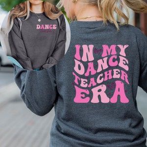 May include: A dark grey long-sleeved sweatshirt with the text "Dance Teacher" in pink on the front and "In My Dance Teacher Era" in pink with butterfly accents on the back.