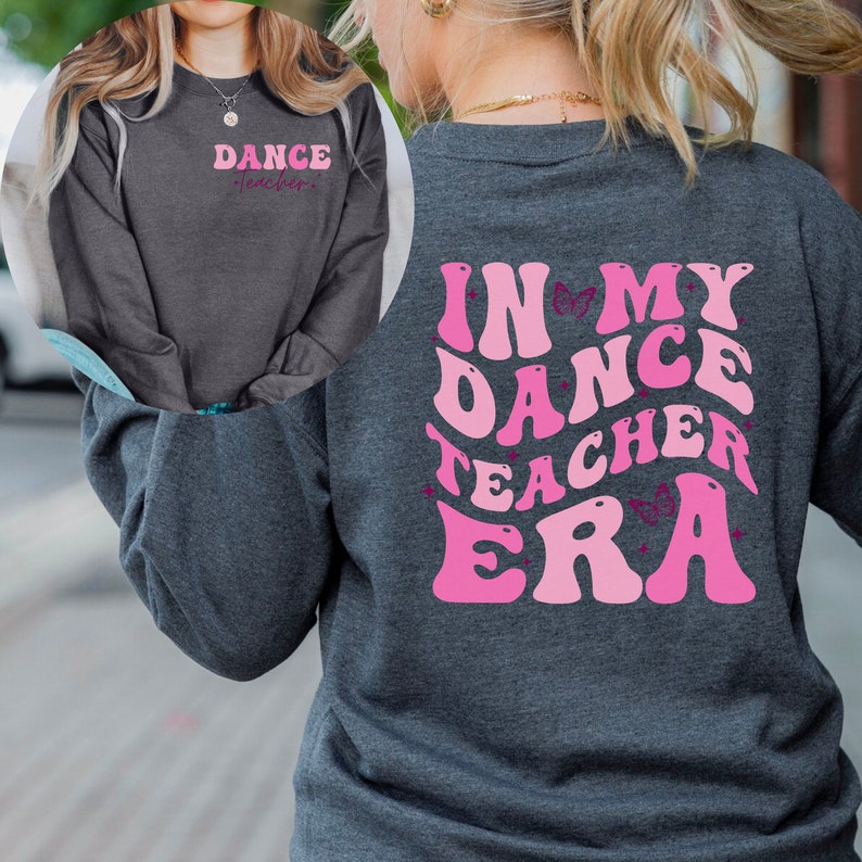 May include: A dark grey long-sleeved sweatshirt with the text "Dance Teacher" in pink on the front and "In My Dance Teacher Era" in pink with butterfly accents on the back.