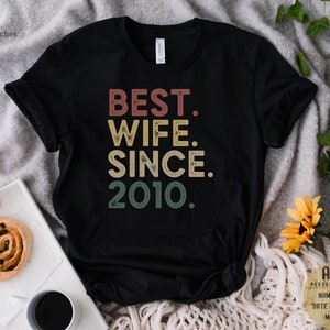 Puede incluir: Camiseta negra con el texto "BEST. WIFE. SINCE. 2010." en una fuente de estilo vintage con colores rojo, naranja, amarillo y verde.