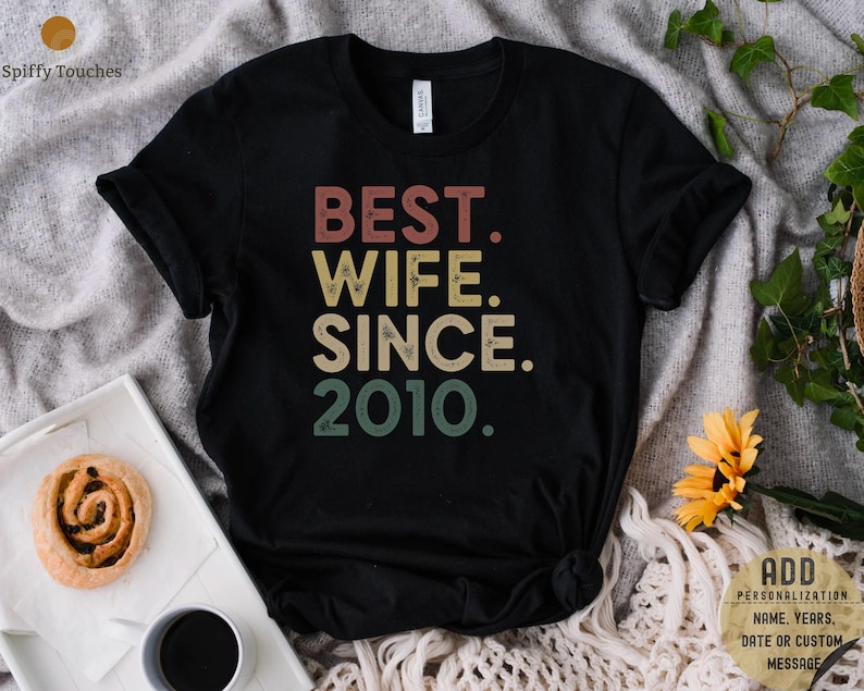Puede incluir: Camiseta negra con el texto "BEST. WIFE. SINCE. 2010." en una fuente de estilo vintage con colores rojo, naranja, amarillo y verde.