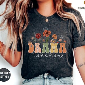 May include: A dark grey heather t-shirt with a retro-style floral design and the text "DRAMA teacher" in colorful letters.