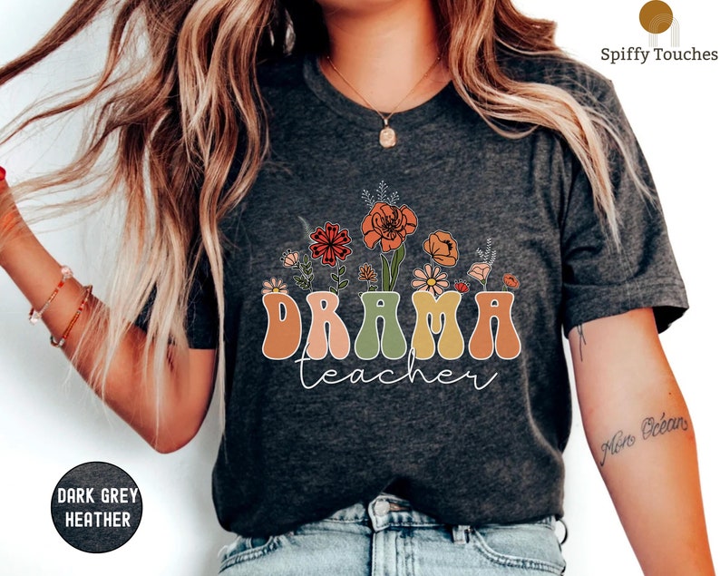 May include: A dark grey heather t-shirt with a retro-style floral design and the text "DRAMA teacher" in colorful letters.