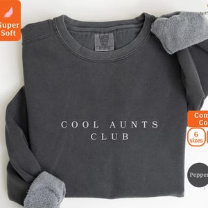 May include: A dark gray sweatshirt with the text "COOL AUNTS CLUB" in white. The sweatshirt has a crew neck and long sleeves with gray fleece cuffs. The image also includes the words "Super Soft" and "Comfort Colors".
