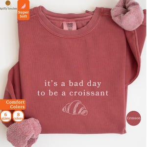 Pode incluir: Camisola de cor carmesim com a frase "it's a bad day to be a croissant" em branco e um desenho de croissant branco. A parte superior de manga comprida tem gola redonda. Uma scrunchie a condizer está na manga.
