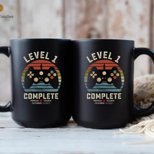 May include: Two black coffee mugs with a retro video game controller graphic and the text "Level 1 Complete". The mugs are personalized with the names "Pamela" and "Steven" and the date "October 19, 2021".