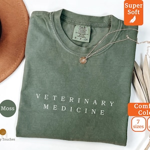 May include: A moss green t-shirt with the words "VETERINARY MEDICINE" printed in white. The shirt has a crew neck and short sleeves. A gold necklace with a pendant is on the shirt. The shirt is labeled "Comfort Colors" and "Super Soft".