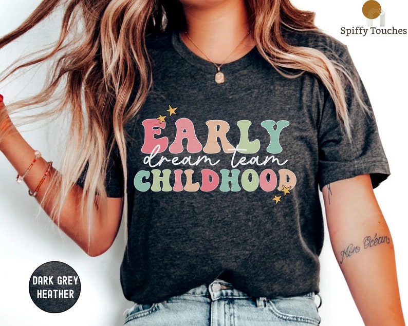 Puede incluir: Una camiseta de color gris oscuro jaspeado con el texto "Early dream team childhood" en letras de colores de estilo retro. Dos peque&ntilde;as estrellas amarillas est&aacute;n a cada lado del texto.