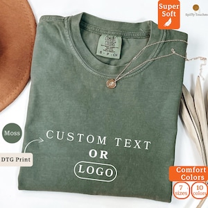 May include: A moss green crew neck t-shirt featuring the white text CUSTOM TEXT OR LOGO printed on the front. The shirt has a Comfort Colors brand label at the inner neckline and is displayed with a delicate gold-toned chain necklace.
