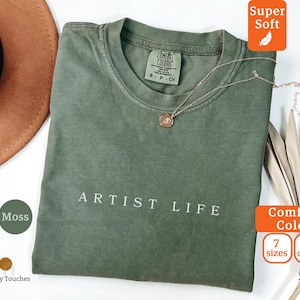 以下が含まれることがあります： 「ARTIST LIFE」の文字が白でプリントされたモスグリーンのTシャツ。クルーネックと半袖です。ペンダント付きのゴールドネックレスがTシャツにかけられています。「Comfort Colors」と「Super Soft」のラベルが付いています。