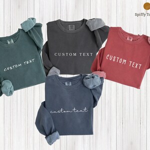 May include: Four long-sleeved sweatshirts in teal, charcoal, red, and navy blue, each with "CUSTOM TEXT" printed in white. The cuffs are folded up, revealing a lighter fabric. The sweatshirts are displayed on a light wood-grain background.